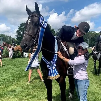 Rider and handler pose with a dark horse wearing blue championship ribbons