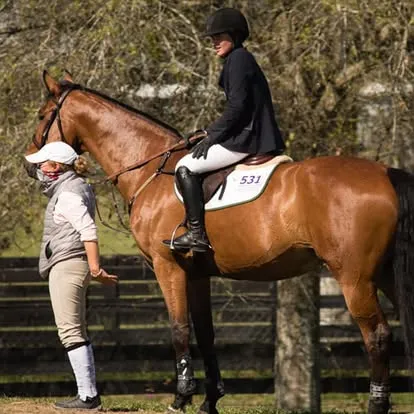 Coach walks alongside a rider on a bay horse near paddock fencing