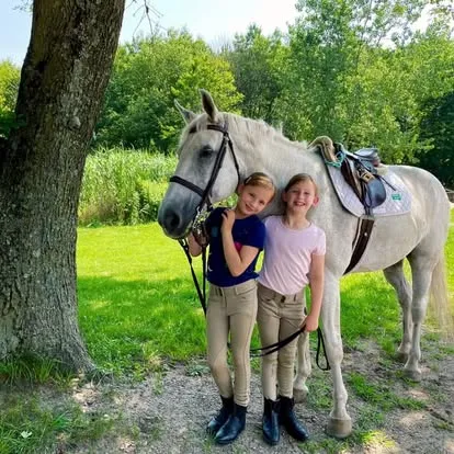 Two smiling young riders standing beside a saddled white pony